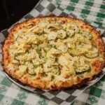 This photo shows a pizza topped with pickles, onions, chives, a cream sauce and ridged potato chips.