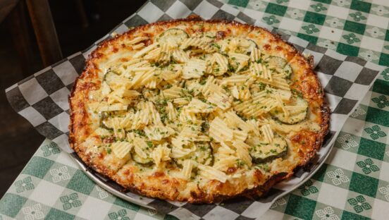 This photo shows a pizza topped with pickles, onions, chives, a cream sauce and ridged potato chips.
