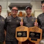 This photo shows contest winner Zac Calder holding his two pizza peel trophies and standing with the competition judges.