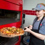 This photo shows a young woman in a Domino's uniform pulling a pizza from the oven.