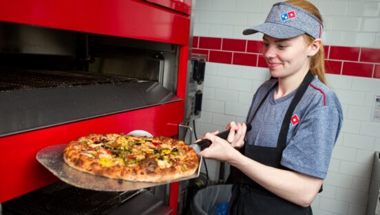 This photo shows a young woman in a Domino's uniform pulling a pizza from the oven.