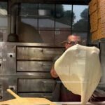 This photo shows a man, viewed through a glass window, stretching dough in his pizza restaurant.