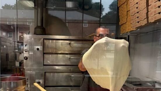 This photo shows a man, viewed through a glass window, stretching dough in his pizza restaurant.