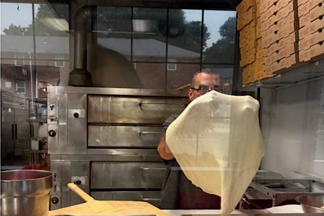 This photo shows a man, viewed through a glass window, stretching dough in his pizza restaurant.
