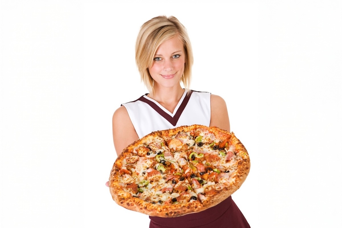 This photo shows a young woman in a cheerleader outfit holding a large pizza with various toppings.