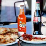 This photo shows several slices of pizza on paper plates with a bottle of Orange Crush and a bottle of Coke.