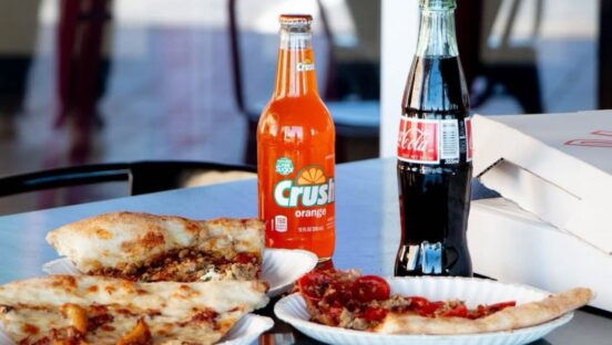 This photo shows several slices of pizza on paper plates with a bottle of Orange Crush and a bottle of Coke.