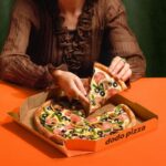 This photo shows a woman, depicted from the neck down, removing a slice of pizza from a whole pie placed on an orange table.