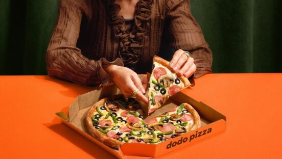This photo shows a woman, depicted from the neck down, removing a slice of pizza from a whole pie placed on an orange table.