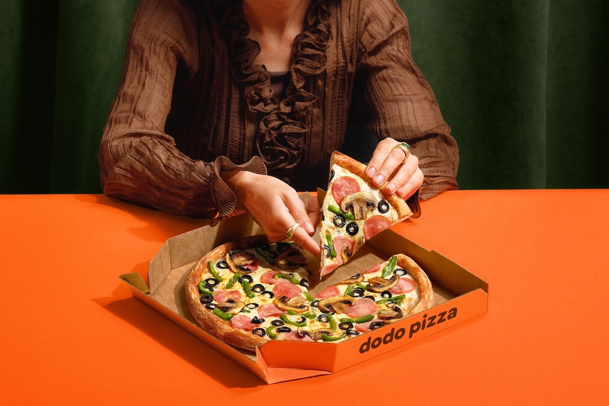 This photo shows a woman, depicted from the neck down, removing a slice of pizza from a whole pie placed on an orange table.