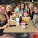 This photo shows a group of five young women seated at a table in a Your Pie location with slices of pizza and drinks in front of them.