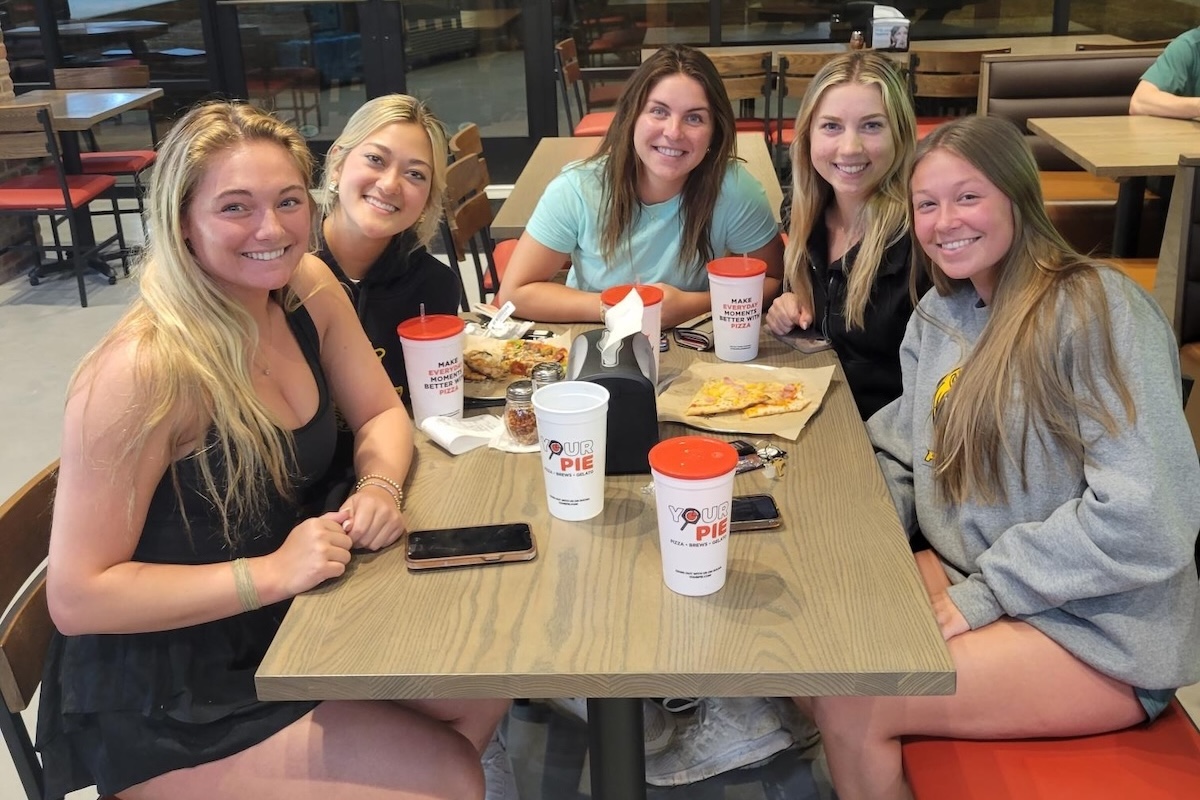 This photo shows a group of five young women seated at a table in a Your Pie location with slices of pizza and drinks in front of them.