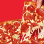 This photo shows a slice being removed from a whole pepperoni pizza with a nice cheese pull. It's set against a red background.