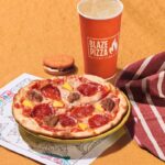 This photo shows a 7" kids pizza, a beverage cup with the Blaze Pizza logo, a cookie and a coloring sheet, with a red and white striped napkin.