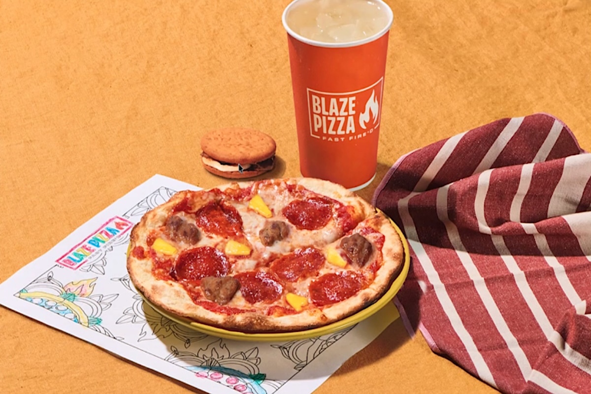 This photo shows a 7" kids pizza, a beverage cup with the Blaze Pizza logo, a cookie and a coloring sheet, with a red and white striped napkin.