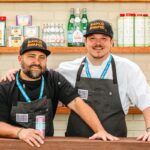 This photo shows Brad Kilgore and Brad Daniels standing in a kitchen, with the latter's arm around the former as they smile for the camera.