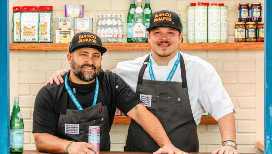 This photo shows Brad Kilgore and Brad Daniels standing in a kitchen, with the latter's arm around the former as they smile for the camera.
