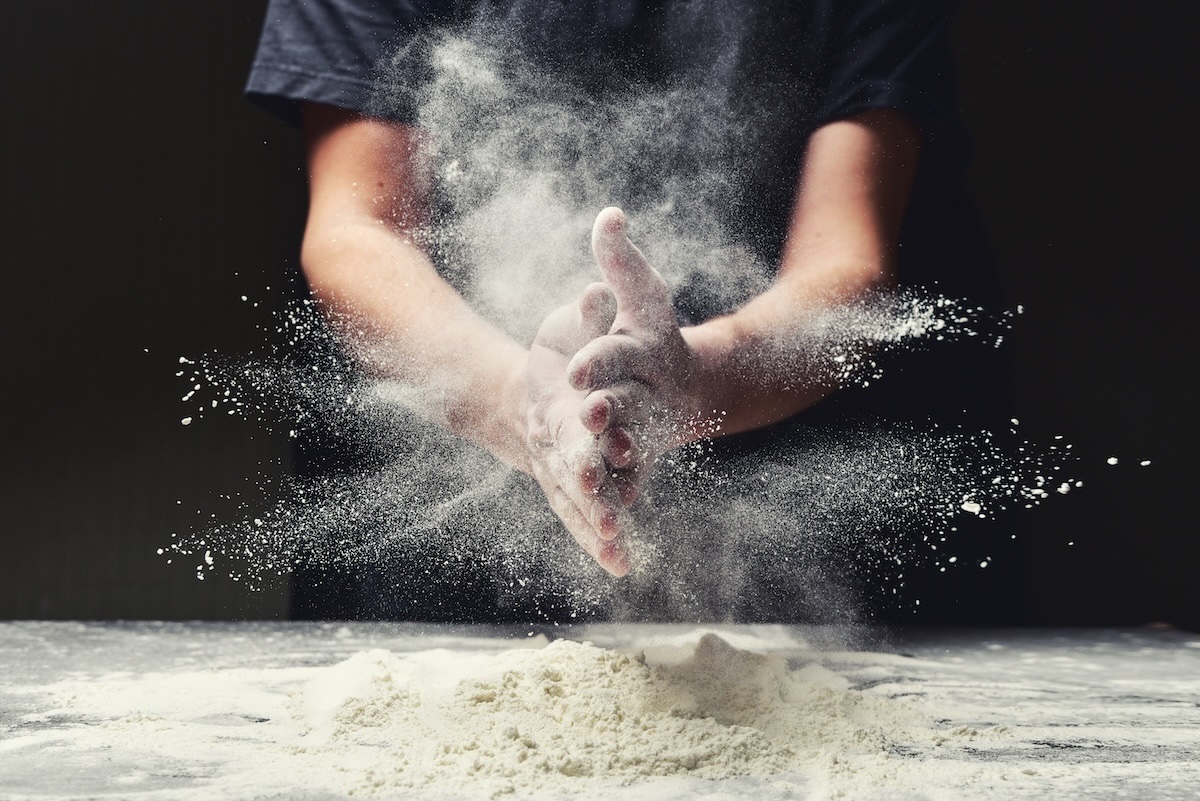 This photo shows a pizza maker clapping his hands together and scattering flour over the make table.