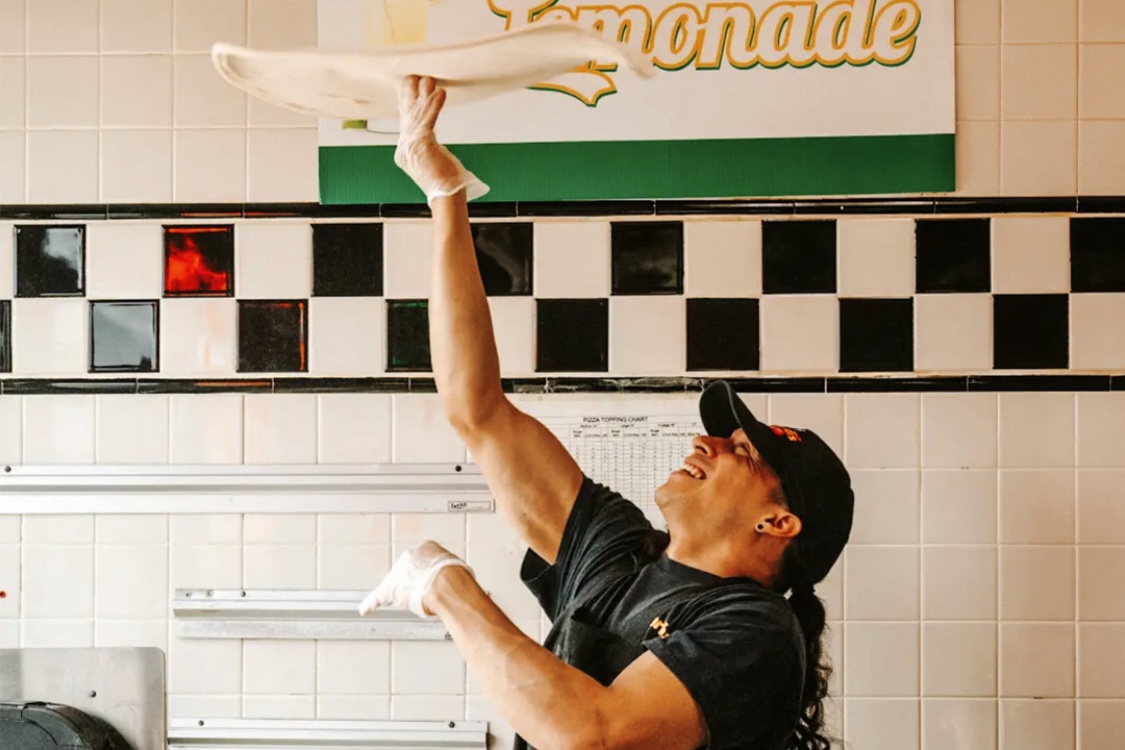 This photo shows a man spinning a round disc of dough high above his head.