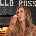 This screenshot shows Katie Lee sitting in front of the pizza oven at one of her restaurants and placing a slice of pizza in her mouth.