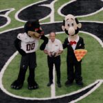 This photo shows Mike LaRosa standing in the middle of the stadium's football field with the Cincinnati Bearcats' mascot and his own brand's mascot, Luigi, all three of them looking up at the camera.