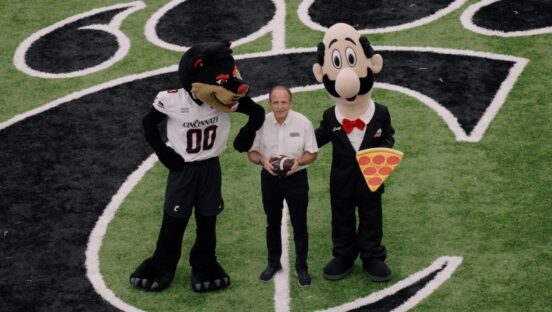 This photo shows Mike LaRosa standing in the middle of the stadium's football field with the Cincinnati Bearcats' mascot and his own brand's mascot, Luigi, all three of them looking up at the camera.