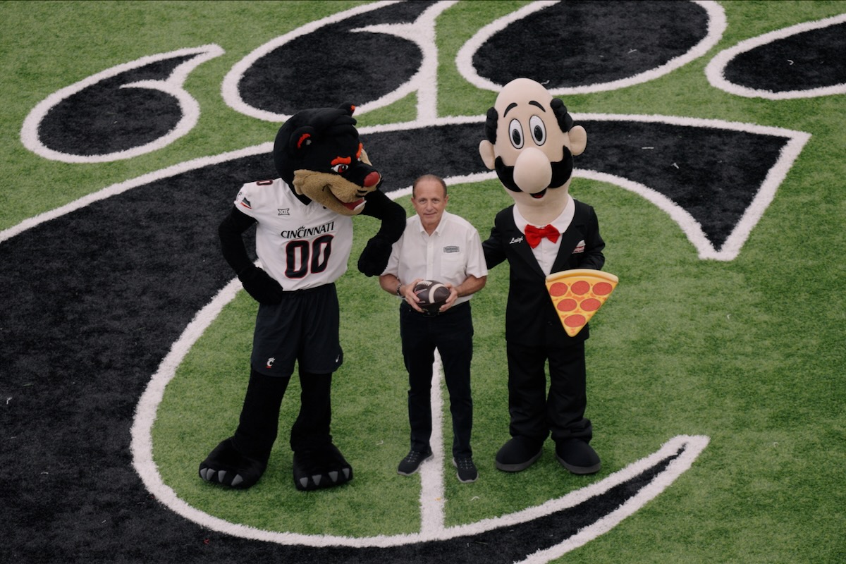 This photo shows Mike LaRosa standing in the middle of the stadium's football field with the Cincinnati Bearcats' mascot and his own brand's mascot, Luigi, all three of them looking up at the camera.