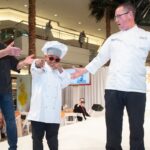 This photo shows a young boy in a chef hat and coat and sunglasses posing coolly and pointing at the camera while two men stand on the stage with him.