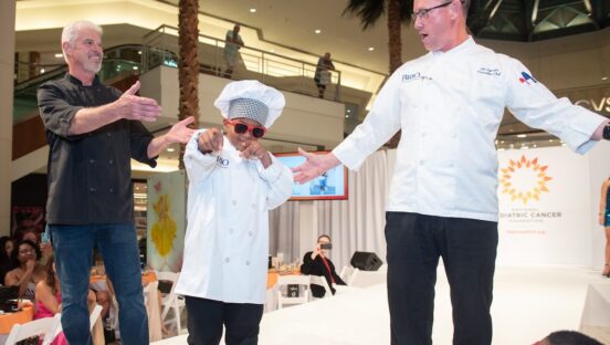 This photo shows a young boy in a chef hat and coat and sunglasses posing coolly and pointing at the camera while two men stand on the stage with him.