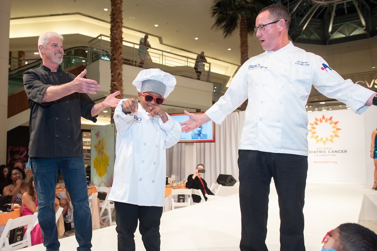 This photo shows a young boy in a chef hat and coat and sunglasses posing coolly and pointing at the camera while two men stand on the stage with him.