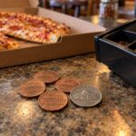 This photo shows four pennies and a nickel next to an open cash register, with a boxed pizza nearby.