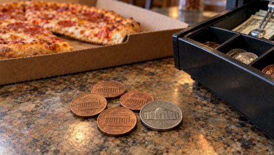 This photo shows four pennies and a nickel next to an open cash register, with a boxed pizza nearby.