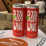This photo shows two cans of Pizza Wine (red with white lettering) sitting on a Modern Apizza pizza box.
