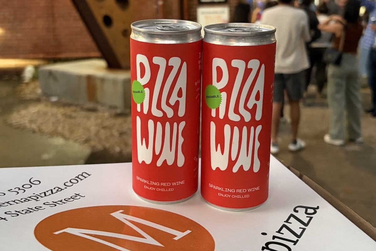 This photo shows two cans of Pizza Wine (red with white lettering) sitting on a Modern Apizza pizza box.