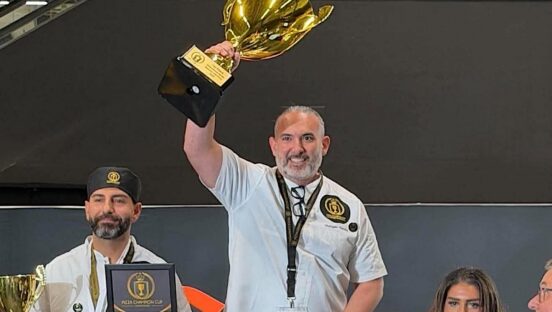 This photo shows Vito Recchia, wearing a white chef coat, holding his trophy in the air at the Pizza Champion Cup International.