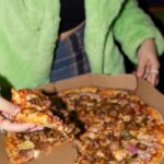 This photo shows a woman in a green jacket pulling a slice from a whole Marco's Pizza pie.
