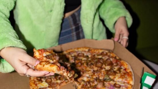 This photo shows a woman in a green jacket pulling a slice from a whole Marco's Pizza pie.