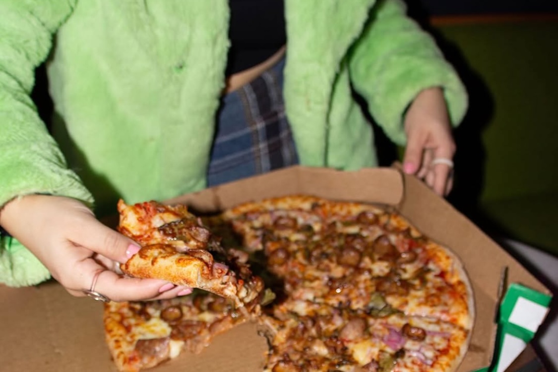 This photo shows a woman in a green jacket pulling a slice from a whole Marco's Pizza pie.