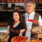 This photo shows Dana Hokin and Robert Garvey, each holding pizzas, with more pizzas on the table in front of them. Robert is wearing his 50 Top Pizza chef coat.