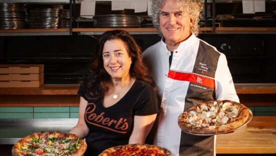 This photo shows Dana Hokin and Robert Garvey, each holding pizzas, with more pizzas on the table in front of them. Robert is wearing his 50 Top Pizza chef coat.