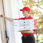 This photo shows a young pizza delivery man in a red uniform and hat ringing the doorbell of a house.