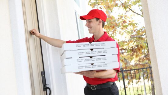 This photo shows a young pizza delivery man in a red uniform and hat ringing the doorbell of a house.