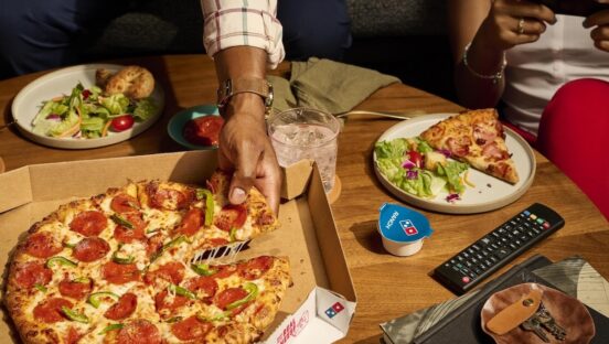 This photo shows a man's hand removing a slice from a whole Domino's pizza.