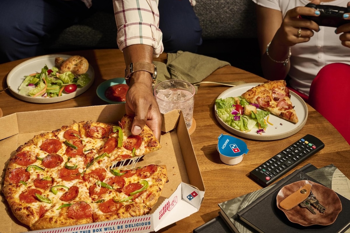 This photo shows a man's hand removing a slice from a whole Domino's pizza.