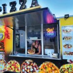 This photo shows Melina Piroso peering out of the window of her mobile pizzeria and holding up a whole pizza.