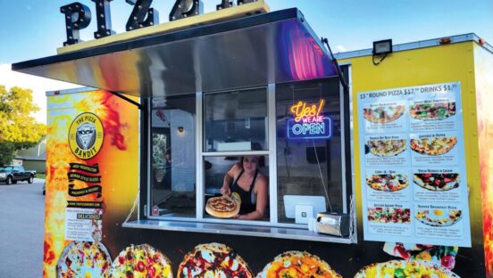 This photo shows Melina Piroso peering out of the window of her mobile pizzeria and holding up a whole pizza.