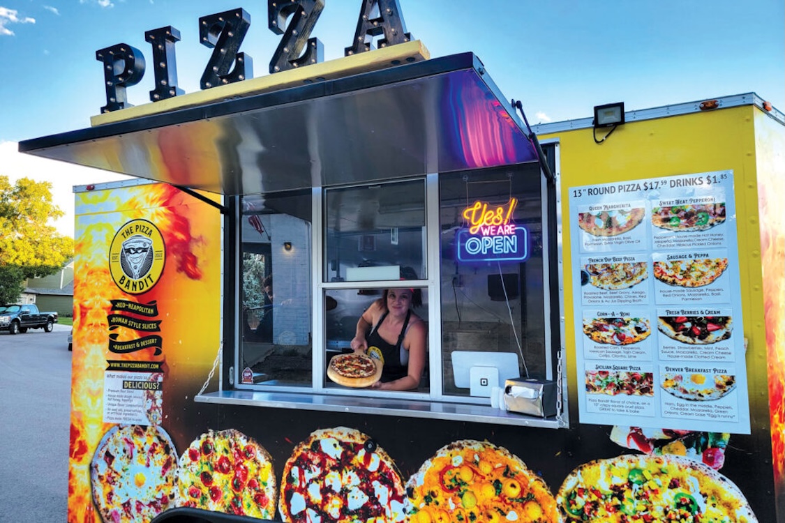 This photo shows Melina Piroso peering out of the window of her mobile pizzeria and holding up a whole pizza.