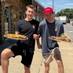 This photo shows two young men, arm in arm, raising their legs in the air in a fun way and each one holding a pizza in front of Mother Bear's Pizza.