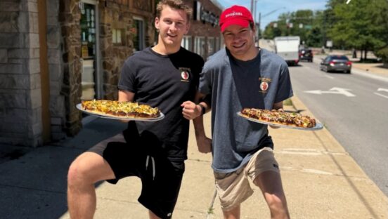 This photo shows two young men, arm in arm, raising their legs in the air in a fun way and each one holding a pizza in front of Mother Bear's Pizza.