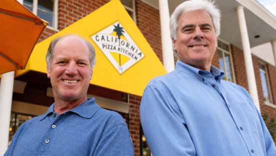 This photo shows Rick Rosenfield and Larry Flax (both wearing dressy blue shirts) standing outside in front of a California Pizza Kitchen sign.